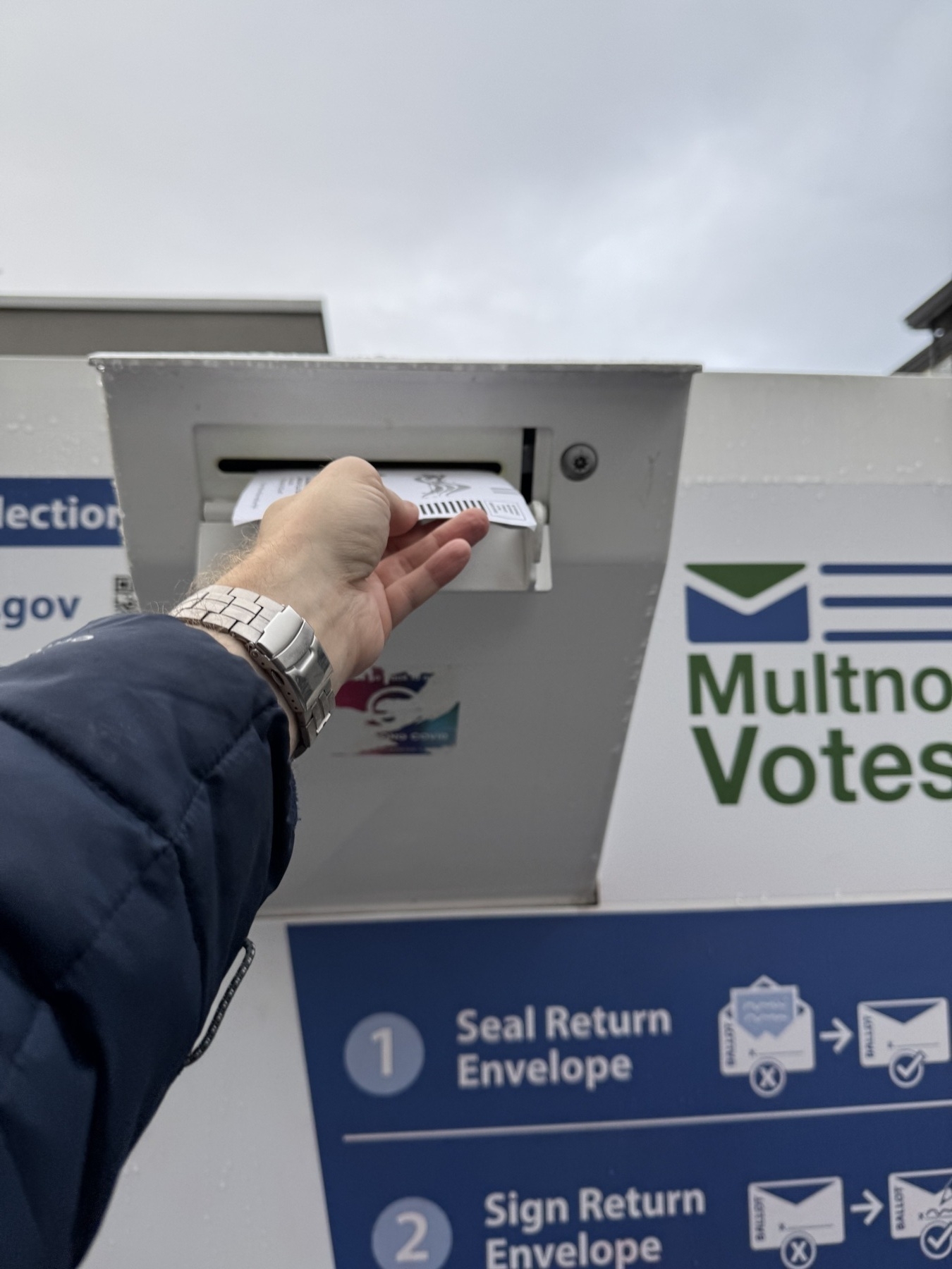 A person is dropping a ballot envelope into an official drop box labeled Multnomah Votes.