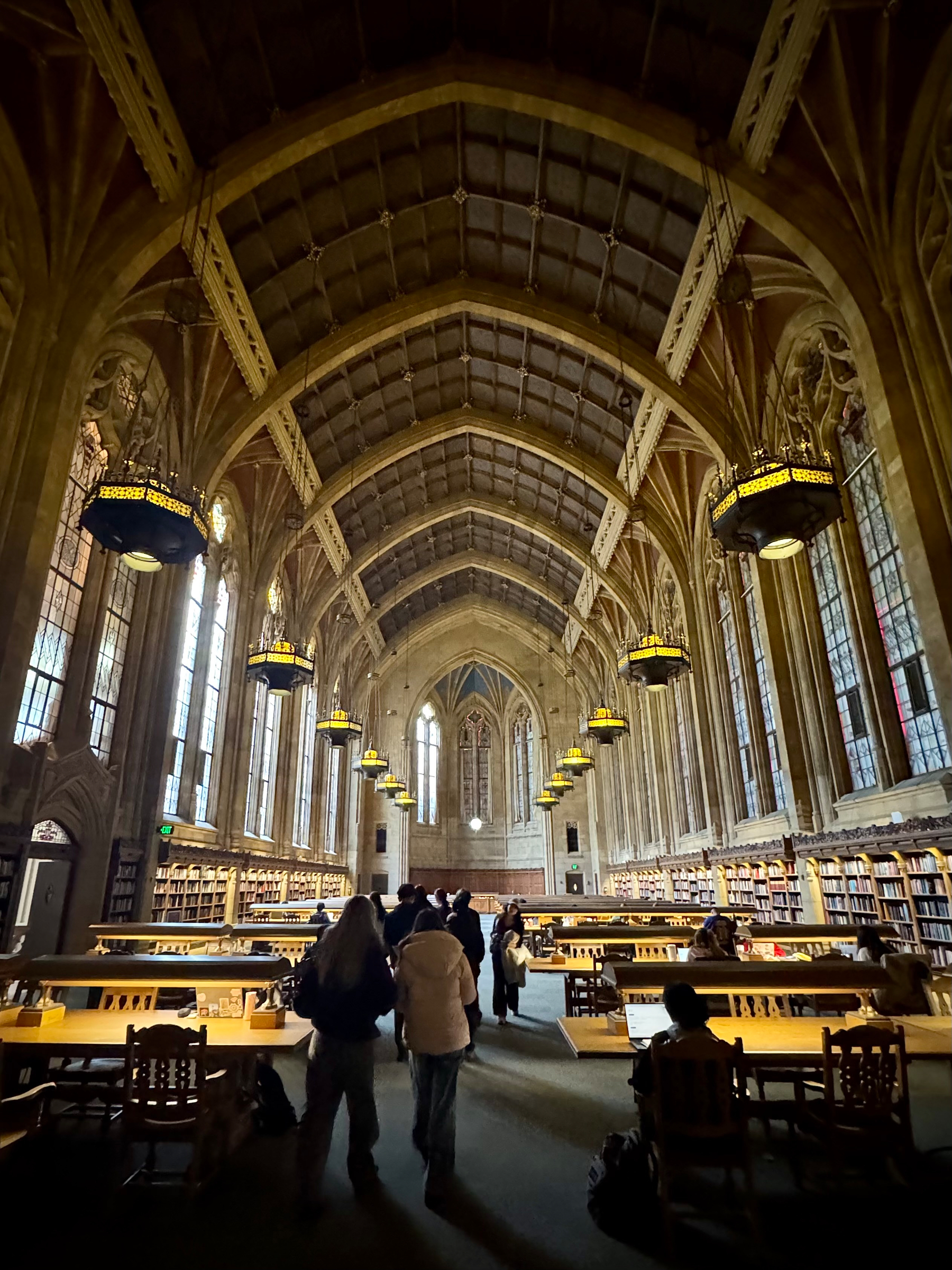 Interior of the Suzzallo Library Reading Room at the University of Washington. The photo looks down a long central aisle beneath a soaring vaulted ceiling with repeating Gothic arches. Warm chandeliers hang in rows, glowing over wooden study tables and tall bookshelves. Large stained-glass windows line the walls, filling the vast, cathedral-like space with soft light. Several people walk toward the far end of the hall, giving a sense of scale to the room’s grand architecture.